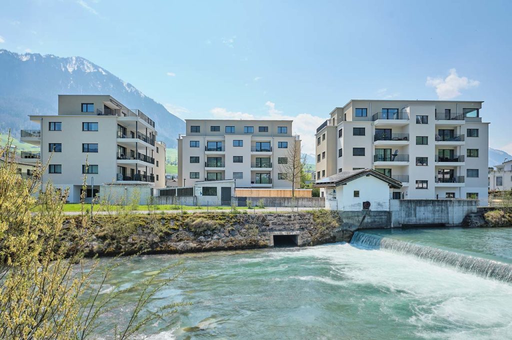 three apartment buildings on the edge of a flowing river with blue sky and mountain peaks behind